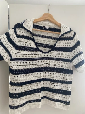Randig virkad topp från H&M - Säljer en kortärmad virkad topp från H&M med breda marinblå och vita ränder. Toppen har djup v-ringning, krage och ett luftigt, hålmönstrat stickat material i bomull. Perfekt för sommaren och ger en avslappnad, trendig look.
