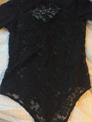 Svart spetsbody med lång ärm - Snygg svart body i genombruten spets med lång ärm och rundad hals. Hela bodyn är täckt av ett detaljerat blommigt spetsmönster som ger en elegant och trendig look. Perfekt att styla med jeans eller kjol för en cool outfit.