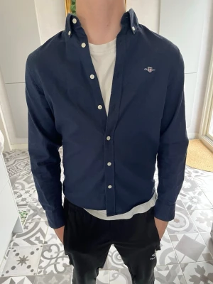 Marinblå skjorta från Gant - Säljer en klassisk marinblå skjorta från Gant med button-down krage och vita knappar. Skjortan har lång ärm och diskret logga broderad på bröstet. Perfekt för dig som gillar stilrena plagg med snygg passform.