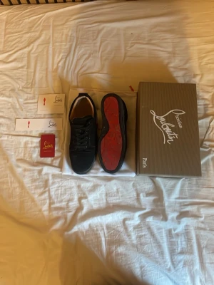 Svarta sneakers från Christian Louboutin - Snygga svarta sneakers från Christian Louboutin med klassisk röd sula och ovandel i mocka. Skorna har lågt skaft, svarta skosnören och diskreta detaljer. Perfekta för dig som vill ha en stilren och exklusiv look.