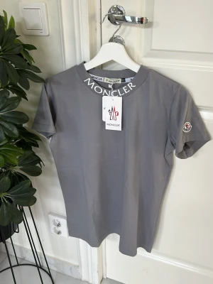 Grå Moncler t-shirt med logga - Snygg grå t-shirt från Moncler med vit logga tryckt på kragen och längs ryggen. Klassisk passform med korta ärmar och rund hals. Tillverkad i mjuk bomull som känns skön mot huden. Perfekt för en clean och stilren look.