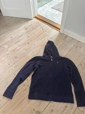 Mörkblå zip hoodie från Gant - Mörkblå hoodie från Gant med dragkedja framtill och två fickor. Klassisk huva och diskret Gant-logga på bröstet. Tillverkad i mjukt material som känns skönt mot huden. Perfekt för en avslappnad och stilren look. skiv för fler bilder😊