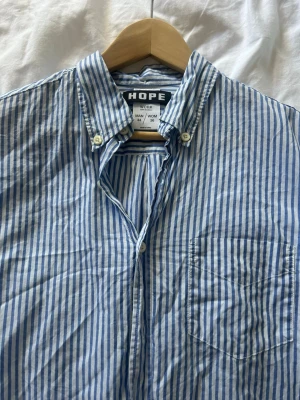 Blårandig skjorta från HOPE - Säljer en blårandig skjorta från HOPE med klassisk button-down krage och bröstficka. Skjortan är tillverkad i bomull och har vita knappar framtill. Perfekt för en clean och avslappnad stil. Passar storlek S