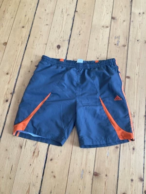 Grå-orange Adidas träningsshorts - Snygga blå träningsshorts från Adidas med orangea detaljer och logga på vänster ben. Shortsen har elastisk midja och är tillverkade i ett lätt och snabbtorkande material, perfekta för sport och aktivitet. Dock är det hål i båda fickorna men det är ett nät runt så att det går att lägga in saker utan att det ramlar ut!!!!