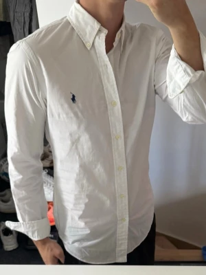 Vit skjorta från Ralph Lauren - Klassisk vit skjorta från Ralph Lauren med button-down krage och broderad marinblå logga på bröstet. Skjortan har lång ärm, knappar framtill och är tillverkad i bomull. Perfekt för en clean och stilren look. 