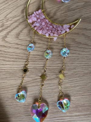 Solfångare med Måne och hjärta prismamobil i guld - Unik dekoration med halvmåne i guldmetall fylld med rosa stenar och hängande kedjor med regnbågsskimrande prismor i hjärtform och solsymboler. Perfekt som smyckesdekoration eller fönsterprydnad för magisk känsla.