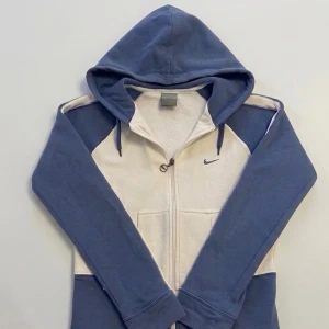 Nike Air hoodie blå och vit  - Snygg Nike Air hoodie i blått och vitt med dragkedja framtill och stor huva. Hoodien har Nike-logga på bröstet och stor Nike Air-tryck på ryggen. Tillverkad i mjukt material med fickor fram och ribbade muddar. Perfekt för en chill och sportig look. Passar kvinnor XS eller barn L 