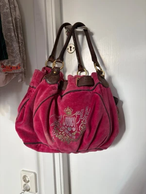 Juicy Couture rosa sammetsväska - Cool rosa handväska från Juicy Couture i mjuk sammet med bruna handtag i läder och guldfärgade metalldetaljer. Väskan har ett glittrigt emblem framtill med krona och text, samt dekorativa sömmar. Perfekt statement piece för dig som gillar färg och bling.