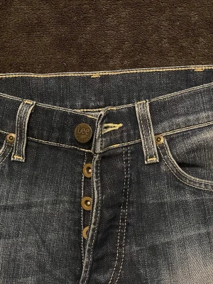 Mörkblå Lee jeans med knappar - Snygga mörkblå jeans från Lee med klassisk knappgylf och kontrastsömmar. Jeansen har fem fickor och tydliga slitningar för en cool, använd look. Tillverkade i denim med Lee-logga på knappen och etiketten i midjan.