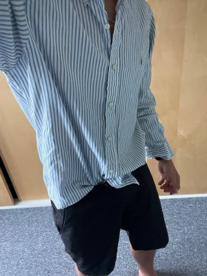 Blåvit randig skjorta från Polo Ralph Lauren - Säljer en klassisk blåvit randig skjorta från Polo Ralph Lauren. Skjortan har långa ärmar, knappar framtill och den ikoniska lilla loggan broderad på bröstet. Tillverkad i mjuk bomull och passar perfekt till jeans eller shorts.