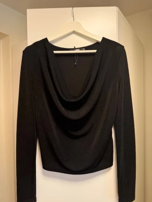 Svart draperad topp från Gina Tricot  - Svart långärmad topp från Gina Tricot med snygg draperad urringning framtill. Toppens material har en lätt glansig finish och sitter figurnära. Perfekt för dig som gillar stilrena och trendiga plagg med det lilla extra.