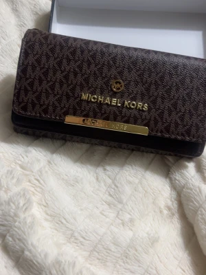 Brun plånbok från Michael Kors - Snygg brun plånbok från Michael Kors med guldfärgade detaljer och logotyp framtill. Plånboken har ett klassiskt MK-mönster och är tillverkad i tåligt konstläder. Perfekt för dig som vill ha en stilren accessoar med plats för kort och kontanter.