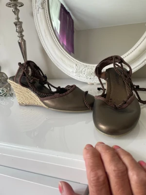 Bruna kilklackade sandaler Gyllene Gripen - Snygga bruna sandaler från Gyllene Gripen med kilklack i flätad jute och snörning runt ankeln. Ovandelen är i brunt tyg och läderimitation, vilket ger en cool och somrig vibe. Perfekta till jeans eller klänning för en avslappnad men trendig look.