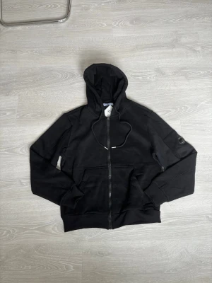 Svart zip hoodie från C.P. Company - Snygg svart hoodie med dragkedja från C.P. Company. Klassisk modell med huva och dragsko, två stora fickor fram och ribbade muddar. Märkets ikoniska logga sitter på vänster ärm. Perfekt för en clean och stilren look.