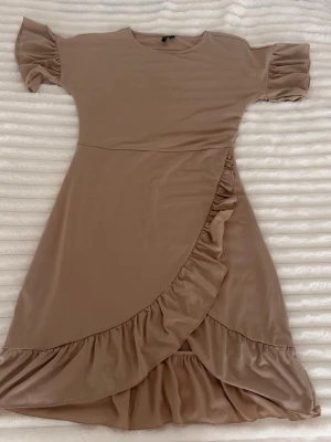 Beige omlottklänning med volang - Supersnygg beige klänning med omlott-design och volangdetaljer längs nederkanten och ärmarna. Klänningen har korta ärmar med rynkade volanger och en mjuk, flowy passform som ger en feminin look. Perfekt för dig som gillar stilrena och trendiga outfits.