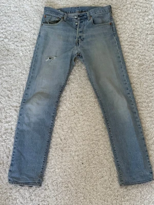 Ljusblå raka Levi's jeans - Säljer ett par klassiska ljusblå jeans från Levi's med rak passform och fem fickor. Jeansen har en liten slitning på vänster lår och är tillverkade i 100% bomull. Perfekta för en avslappnad och trendig look.