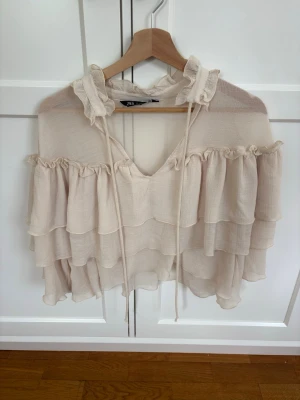 Beige volangblus från Zara - Supersöt beige blus från Zara med flera volanglager och romantiska detaljer. Blusen har en hög krage med volang och knytband samt ett luftigt, transparent tyg som ger en bohemisk känsla. Perfekt till jeans eller kjol för en trendig look.