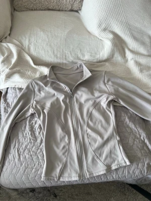Ljusbeige/grå vindjacka med dragkedja - Snygg ljusbeige/grå vindjacka med hel dragkedja framtill och figurnära passform. Jackan har hög krage och långa ärmar, perfekt för lager på lager. Materialet känns mjukt och lätt, vilket gör den enkel att bära till olika outfits.