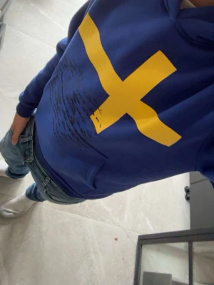 Blå hoodie med gultt kors - Säljer en blå hoodie med ett stort gult kors på bröstet, inspirerat av svenska flaggan. Tröjan kommer från ett helt eget märke som kallas Concrete ink. Skick på tröjan är 10/10 använd en gång skriv vid fundering eller frågor svarar alltid pris går att diskutera/mvh William