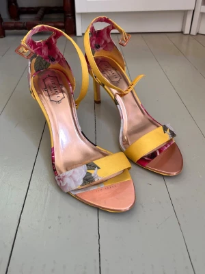 Ted Baker klackar - Säljer ett par färgglada klackskor från Ted Baker med gul rem och blommigt mönster på sidorna. Skorna har öppen tå, smal klack och justerbart spänne runt ankeln. Insidan är metallic-rosa och ger en lyxig känsla. Perfekta för dig som vill sticka ut! Nypris ca. 2000kr