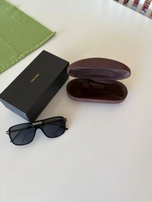 Svarta Tom Ford solglasögon - Snygga svarta solglasögon från Tom Ford med klassisk rektangulär form och mörka glas. Metall- och plastdetaljer ger en exklusiv känsla. Kommer med originalfodral och box. Perfekta för dig som vill ha en stilren accessoar till sommaren.