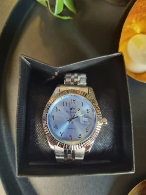 Silverfärgad klocka från Ricecgo - Stilren klocka från Ricecgo med silverfärgat metallarmband och ljusblå urtavla. Urtavlan har arabiska siffror och datumvisning. Klassisk design med räfflad bezel och länkarmband som ger en modern vibe. Helt ny aldrig använd och ej kortad så alla länkar ingår