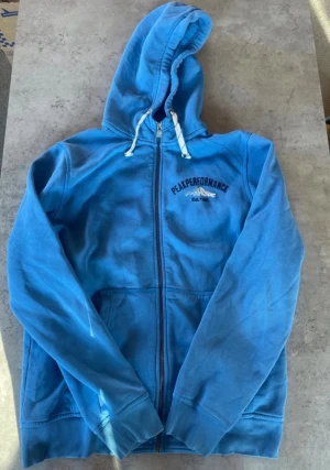 Blå zip hoodie från Peak Performance - Snygg blå hoodie från Peak Performance med dragkedja framtill och vit snörning i huvan. Broderad logga och bergmotiv på bröstet. Tillverkad i mjuk bomullsblandning och har två fickor fram. Perfekt för en avslappnad och sportig stil. Skick syns på bilden inget som syns så mycket på. Storlek xxl