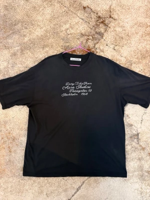  Acne Studios t-shirt - Snygg svart t-shirt från Acne Studios med vit broderad text på bröstet. Klassisk rund halsringning och avslappnad passform. Perfekt för dig som gillar stilrena plagg med en twist.