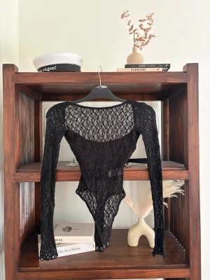 Svart spetsbody med långa ärmar - Säljer en svart body i genombruten spets med långa ärmar och rund halsringning. Bodyn har tryckknappar i grenen och ett elegant, transparent mönster som ger en trendig look. Perfekt för dig som vill ha något extra till outfiten.