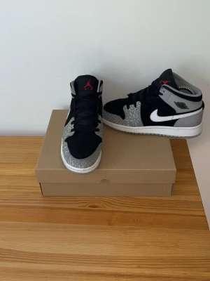 Air Jordan 1 Mid svart/grå/vit - Snygga Air Jordan 1 Mid sneakers i svart, grått och vitt med klassisk elefantmönstrad detalj på tå och sidor. Skorna har svart mocka, vita sulor och röda detaljer på plösen. Ikonisk Swoosh-logga på sidan och Wings-märke vid ankeln.