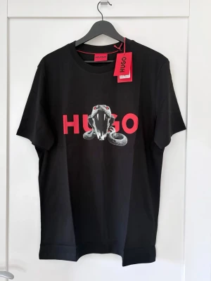 Hugo T-shirt, M, Ny! Ord. Pris 799kr. - Hugo T-shirt med ormgrafik. Normal passform.  Axelbredd: 44 cm Längd: 70 cm Mellan armhålorna: 53 cm 