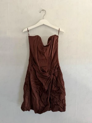 Brun kort klänning med drapering - Snygg brun kort klänning med tunna axelband och draperad detalj framtill. Klänningen har en lätt glansig yta och en unik formad byst. 