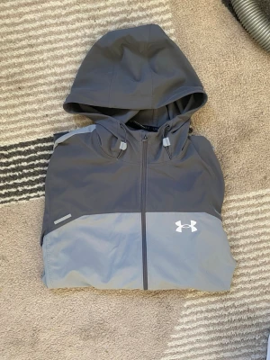 Grå vindjacka från Under Armour - Snygg tvåtonad grå vindjacka från Under Armour med huva och hel dragkedja. Jackan har justerbar huva, två sidofickor med dragkedja och logga på bröstet. Tillverkad i lätt och vattenavvisande material, perfekt för blåsiga dagar.