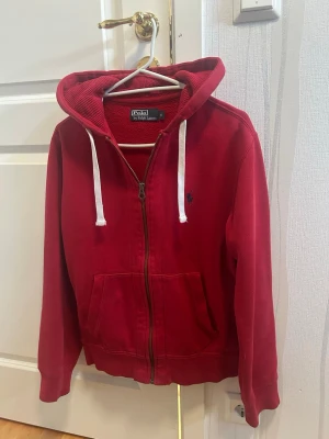 Röd hoodie från Polo Ralph Lauren - Snygg röd hoodie från Polo Ralph Lauren med dragkedja, vita snören i huvan och klassisk blå logga på bröstet. Tillverkad i mjukt bomullsmaterial och har två fickor framtill. Perfekt för en avslappnad och stilren look.