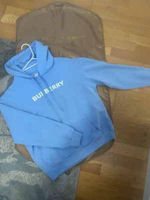 Blå hoodie från Burberry - Snygg ljusblå hoodie från Burberry med vit logga tryckt över bröstet. Klassisk modell med huva och dragsko samt en stor magficka. Perfekt för dig som vill ha en stilren och trendig look. Mjuk bomullskänsla och relaxed fit.
