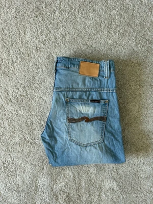 Ljusblåa Nudie Jeans Slim Fit Storlek W34  - Mycket fina Nudie Jeans i blå färg. | Jeansen är i bra skick och sparsamt använda. |  Äkta som allt annat jag säljer! ✅ |  Storlek: W34 L34 | Skick: Bra Skick - bakpatchen har blivit utbytt, lagade på ett ställe och tyvärr lite missfärgning på bakre fickan. | Färg: Blå | Märke: Nudie | Modell: Jeans |   Hör gärna av dig vid frågor så svarar jag så fort jag kan! 😁 |  (SKU - 192)