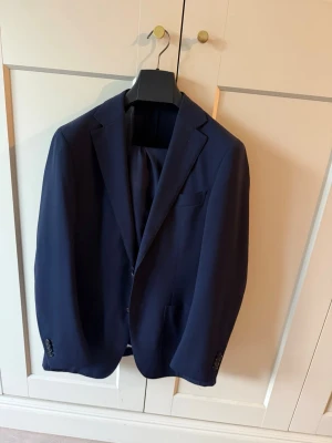 Mörkblå kostym från Suitsupply - Snygg mörkblå kostym från Suitsupply med klassisk kavaj och matchande byxor. Kostymen har två knappar, fickor fram och smal passform. Perfekt för dig som vill ha en stilren och modern look. Byxa size 24, model Soho. Nypris 6500