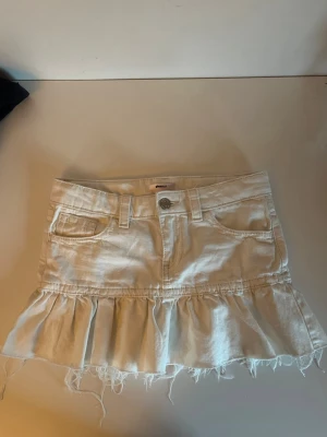 Vit jeans kjol - Supersöt vit jeanskjol med volang och rå, fransig kant. Kjolen har klassiska jeansfickor fram och bak samt bälteshällor. Perfekt för dig som gillar en trendig och avslappnad stil.