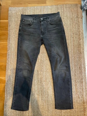 Svarta lean dean jeans från Nudie - Snygga svarta lean dean jeans från Nudie med klassisk femficksmodell och diskreta slitningar. Jeansen har en något tvättad look och Nudie-loggan broderad på bakfickorna. Tillverkade i mjuk och stretchig denim för skön passform.