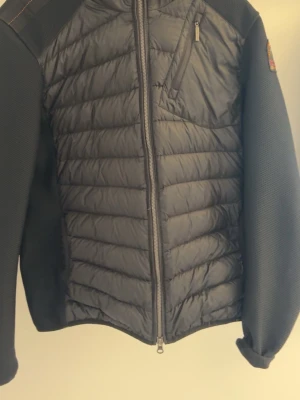 Svart pufferjacka från Parajumpers - Snygg svart pufferjacka från Parajumpers med quiltad front och mesh-liknande ärmar. Jackan har dragkedja framtill, bröstficka med zip och klassisk PJS-patch på ärmen. Perfekt för kalla dagar och riktigt stilren look.