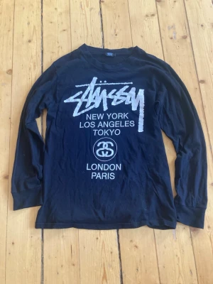Svart långärmad Stüssy t-shirt - Snygg svart långärmad t-shirt från Stüssy med ikoniskt tryck framtill i vitt med städer som New York, Los Angeles, Tokyo, London och Paris. Tillverkad i mjuk bomull och har en avslappnad passform. Perfekt för dig som gillar streetwear och vill sticka ut.