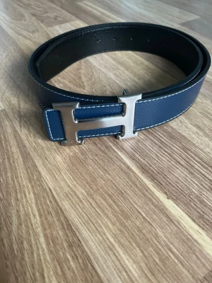 Marinblått H bälte  - Säljer ett marinblått bälte med ett H på. Bältet är 105cm långt och är justerbart. De är i läder material. Skickar snabbt, pris går att diskuteras!🤝📦