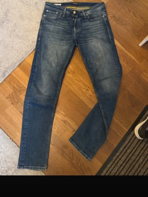 Jack and Jones tapered Mike - Säljer ett par blå tapered jeans från Jack & Jones med snygga slitningar och klassisk femficksmodell. Jeansen har guldfärgade knappar och detaljer samt normal midja. Perfekta för en avslappnad och trendig stil.              W31 L34