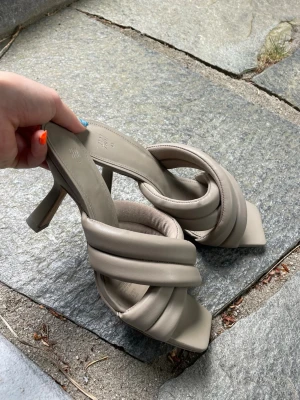 Beige flätade klackade sandaler, strlk 40 snarare - Säljer ett par trendiga beige sandaler med flätade, vadderade remmar och fyrkantig tå. Skorna har en smal klack och öppen häl, vilket ger en modern look. Tillverkade i ett mjukt, läderliknande material. Perfekta för dig som gillar stilrena detaljer.