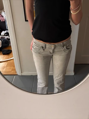 Ljusgrå bootcut jeans - Säljer ett par ljusgrå bootcut jeans med klassiska femficksdetaljer och låg midja. Jeansen har en snygg, avslappnad passform och är tillverkade i denim med lite stretch för extra komfort. Perfekt för dig som gillar en cool och enkel stil.