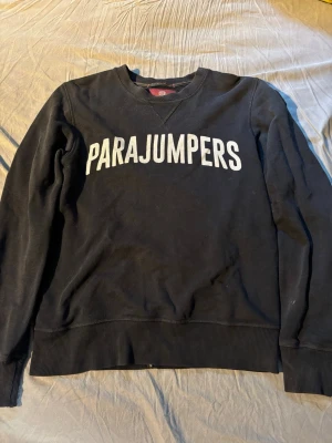 Svart sweatshirt från Parajumpers - Svart sweatshirt från Parajumpers med stor vit logga på bröstet och ribbade muddar vid ärmslut och nederkant. Klassisk rund halsringning och patch på ena ärmen. Tvättar tröjan innan jag skickar☺️