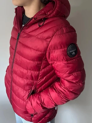 Röd pufferjacka från Napapijri - Snygg röd pufferjacka från Napapijri med huva och svart logomärke på ärmen. Jackan har dragkedja framtill, två sidofickor och quiltad design. Perfekt för kalla dagar och ger en sportig look.