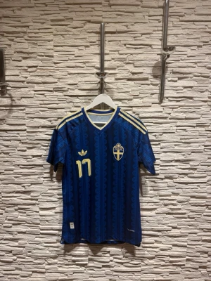 Blå Sverige matchtröja Adidas - Säljer en blå matchtröja från Adidas för Sveriges landslag med gula detaljer, nummer 17 och namnet Gyökeres på ryggen. Tröjan har korta ärmar, V-ringning och klassiska Adidas-ränder på axlarna. Perfekt för fotbollsfans! Priset kan diskuteras!