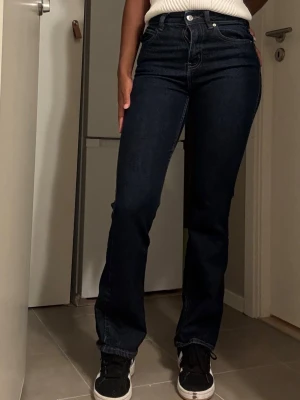 Mörkblå raka jeans med hög midja - Snygga mörkblå jeans med raka ben och hög midja. Klassisk femficksmodell med silverfärgade knappar och nitar. Jeansen har en stilren look och passar perfekt till sneakers eller boots. Materialet är kraftig denim med lite stretch för skön passform.