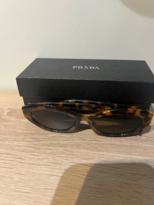 Prada solglasögon i leopardmönster - Snygga solglasögon från Prada med trendigt leopardmönster och mörka glas. Bågarna har en rundad, något cat-eye form och är tillverkade i ett glansigt syntetmaterial. Kommer med originaletui och äkthetskort.